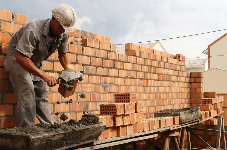 Pedreiro aplica argamassa na construção de uma parede de tijolos, sobre andaime, em obra urbana sob céu parcialmente nublado. Cena retrata o trabalho na construção civil.