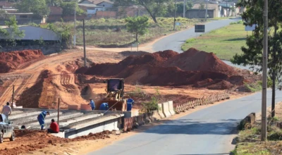 A imagem mostra as obras de duplicação da ponte na Avenida Casemiro de Abreu, com vigas instaladas, operários em atividade e máquinas realizando terraplanagem.