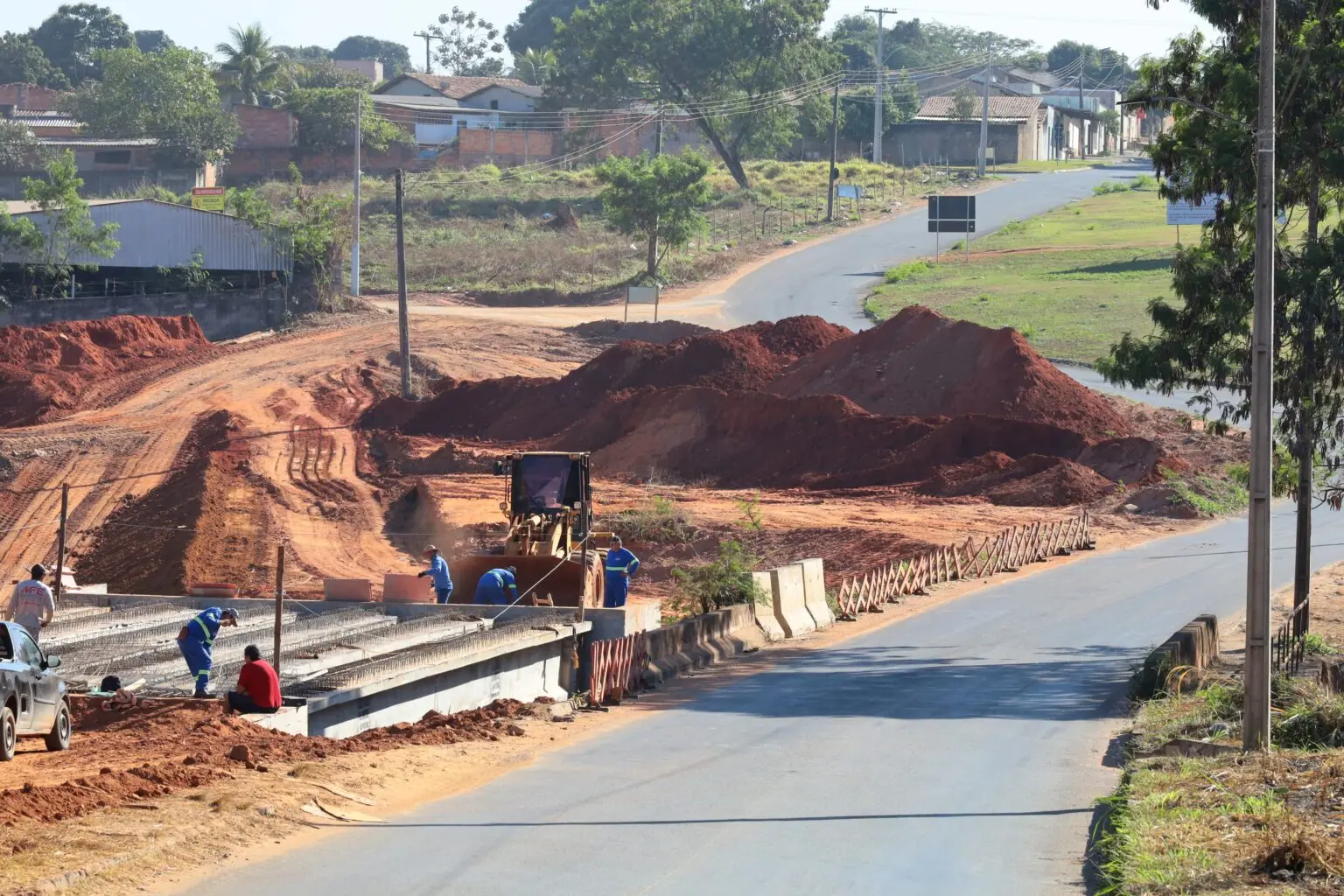 A imagem mostra as obras de duplicação da ponte na Avenida Casemiro de Abreu, com vigas instaladas, operários em atividade e máquinas realizando terraplanagem.