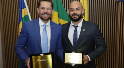 Dois homens de terno, sendo um deles o vereador Lipe Gomes, seguram placas de homenagem, com bandeiras do Brasil, Goiás e Aparecida de Goiânia ao fundo.