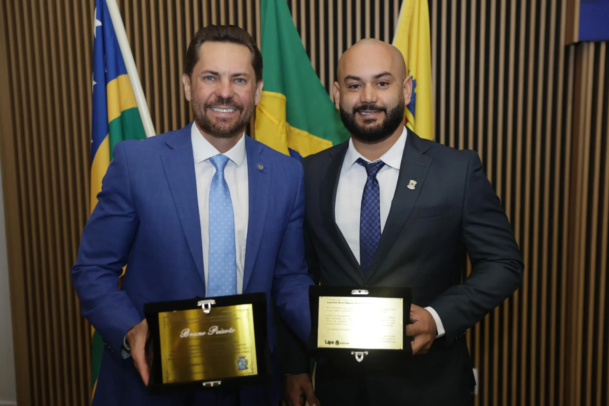 Dois homens de terno, sendo um deles o vereador Lipe Gomes, seguram placas de homenagem, com bandeiras do Brasil, Goiás e Aparecida de Goiânia ao fundo.