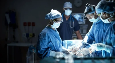 Equipe médica realiza procedimento cirúrgico em ambiente hospitalar, usando aventais, luvas, máscaras e iluminação especializada, enquanto outro profissional acompanha e monitora a operação.