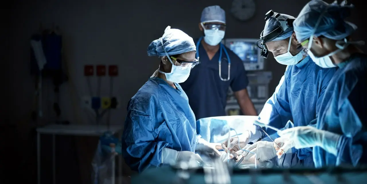 Equipe médica realiza procedimento cirúrgico em ambiente hospitalar, usando aventais, luvas, máscaras e iluminação especializada, enquanto outro profissional acompanha e monitora a operação.