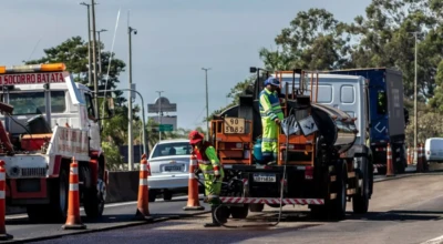 Equipe de manutenção asfáltica recapeia faixa de rodovia urbana com cones de sinalização.