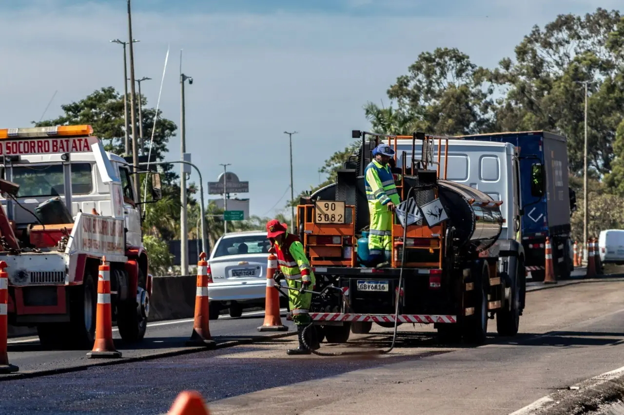 Equipe de manutenção asfáltica recapeia faixa de rodovia urbana com cones de sinalização.