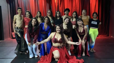 Grupo de bailarinos posa em frente a cortina vermelha, com destaque para dançarina ao centro em traje típico de dança oriental.