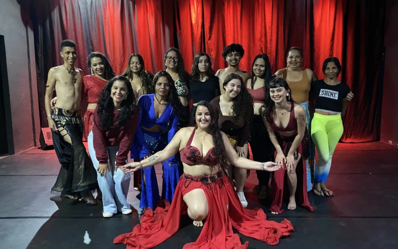 Grupo de bailarinos posa em frente a cortina vermelha, com destaque para dançarina ao centro em traje típico de dança oriental.