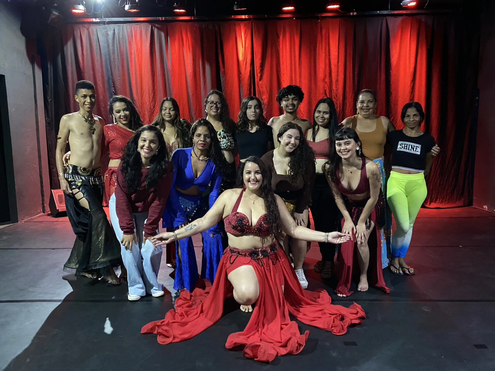 Grupo de bailarinos posa em frente a cortina vermelha, com destaque para dançarina ao centro em traje típico de dança oriental.