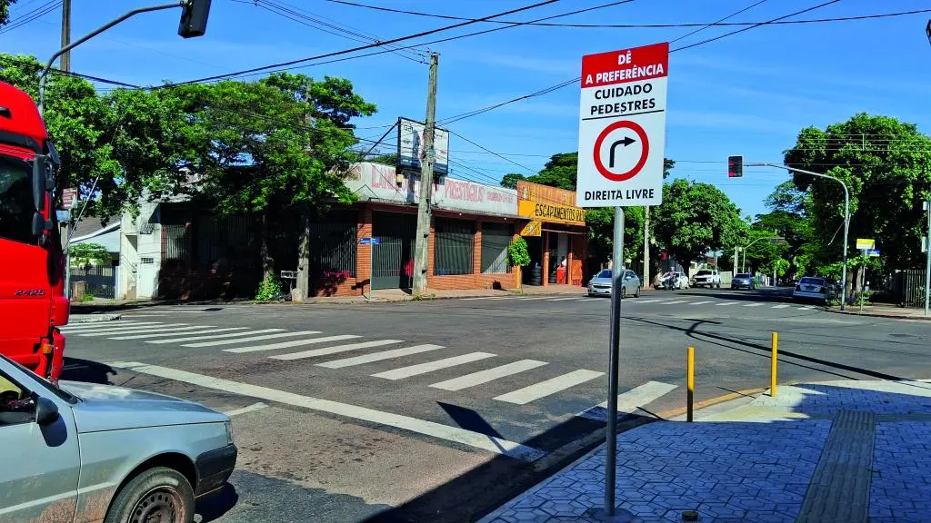 A imagem mostra um cruzamento urbano sinalizado, com faixa de pedestres e placa indicando “Dê a preferência – Cuidado pedestres – Direita livre”.