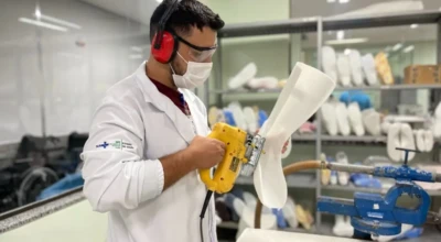 Profissional confecciona órtese com lixadeira elétrica em oficina ortopédica, usando jaleco, máscara e protetores auriculares.
