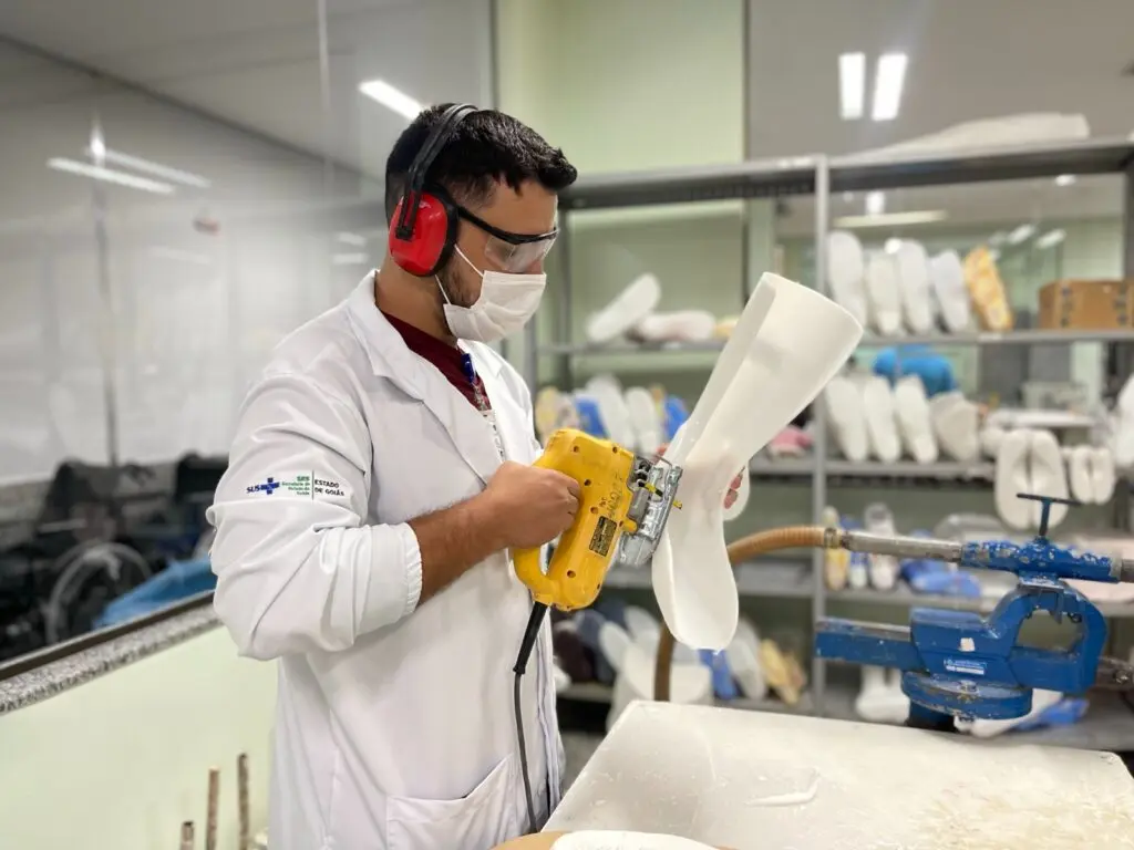 Profissional confecciona órtese com lixadeira elétrica em oficina ortopédica, usando jaleco, máscara e protetores auriculares.