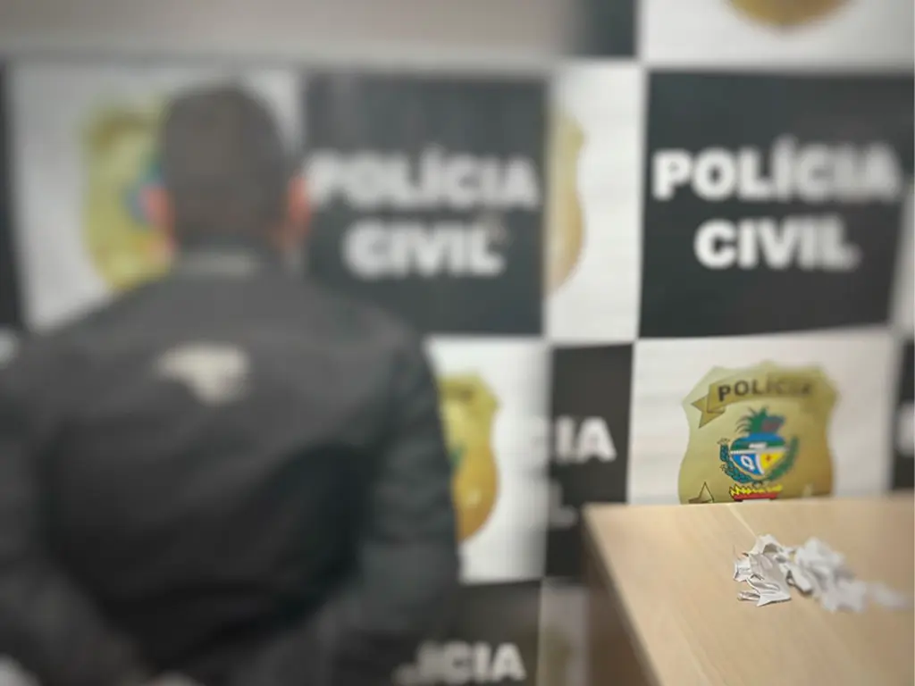 A imagem mostra um homem de costas, com as mãos para trás, diante de um painel da Polícia Civil. Sobre a mesa ao lado, há vários pedaços de papel rasgados, sugerindo documentos destruídos.