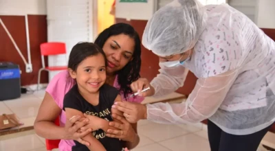 O ChatGPT disse: Criança sorri enquanto recebe vacina no braço, sentada no colo de uma mulher, sob os cuidados de uma profissional de saúde.