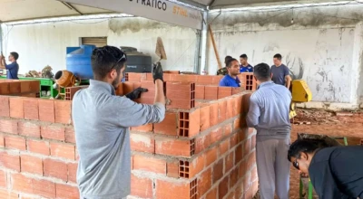 Alunos participam de aula prática de alvenaria, assentando tijolos em um ambiente de treinamento com apoio de ferramentas e orientação técnica.
