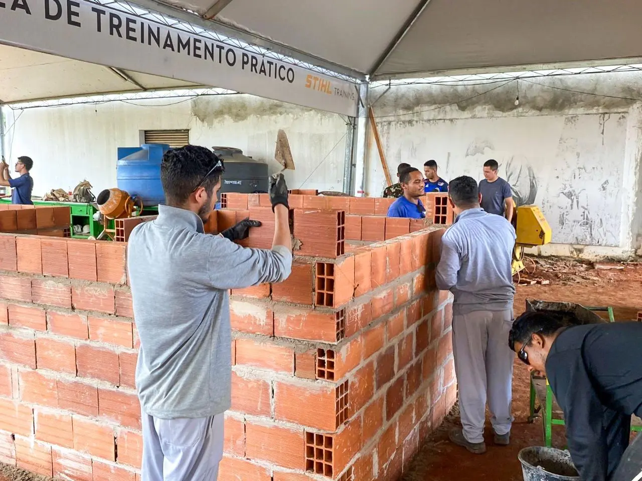 Alunos participam de aula prática de alvenaria, assentando tijolos em um ambiente de treinamento com apoio de ferramentas e orientação técnica.