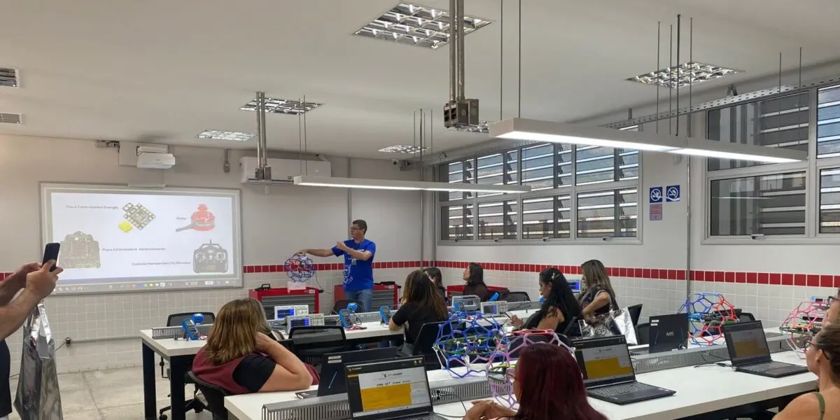 A imagem mostra um instrutor apresentando um modelo geométrico a um grupo em um laboratório moderno, equipado com notebooks e materiais de robótica, durante uma aula prática de tecnologia.