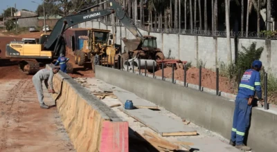 Trabalhadores atuam na construção de uma galeria de concreto, com uso de escavadeiras e tratores, em obra de drenagem em área urbana.