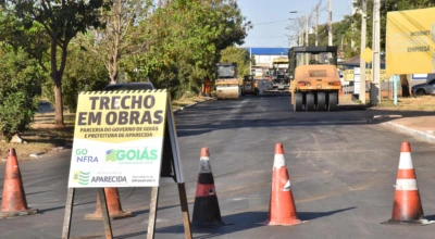 A imagem mostra trecho em obras de recapeamento em Aparecida, com placa de aviso, cones de sinalização e máquinas aplicando novo asfalto.