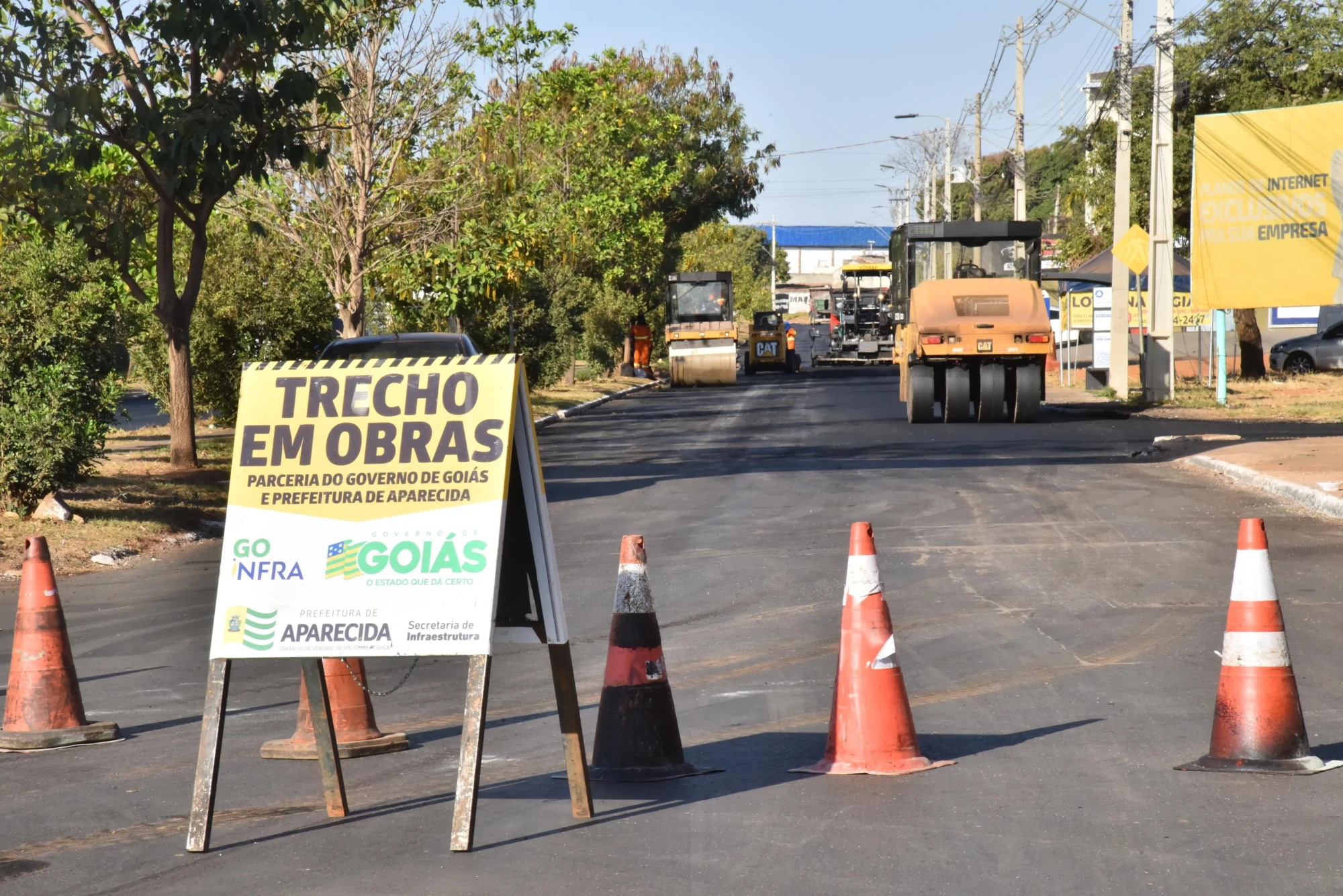 A imagem mostra trecho em obras de recapeamento em Aparecida, com placa de aviso, cones de sinalização e máquinas aplicando novo asfalto.