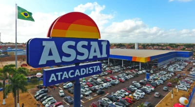 Placa do Assaí Atacadista em destaque, loja ao fundo com estacionamento cheio e bandeira do Brasil ao lado.