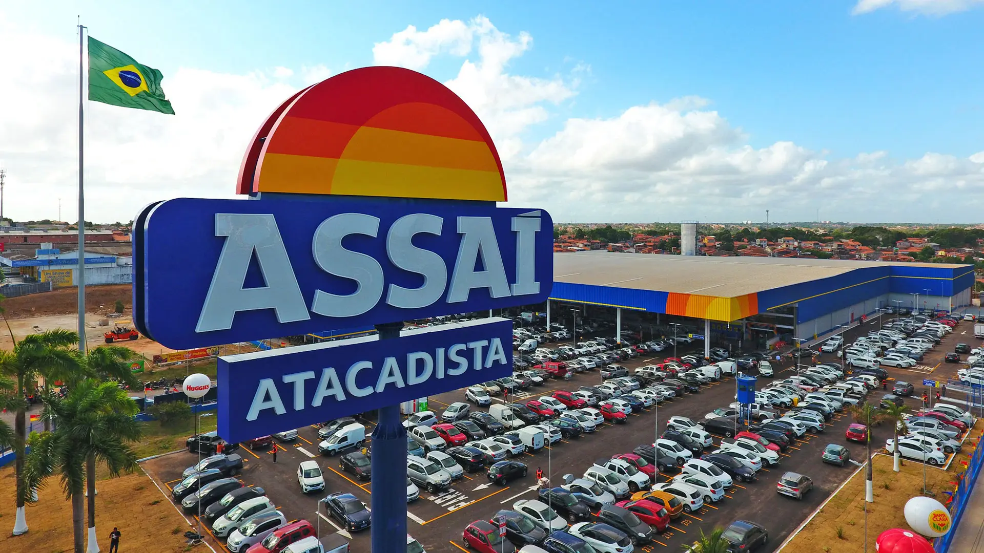Placa do Assaí Atacadista em destaque, loja ao fundo com estacionamento cheio e bandeira do Brasil ao lado.