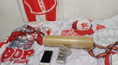 Mesa com bandeira e camisas do Vila Nova F.C., boné vermelho e branco, celular, balança de precisão, invólucro com substância e um tubo de papelão.
