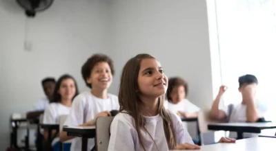Alunos em sala de aula, de camisetas brancas, atentos e sorrindo.