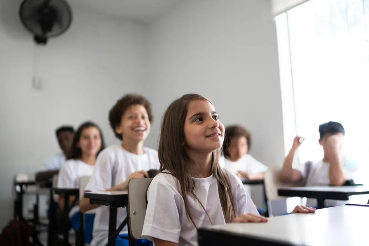 Alunos em sala de aula, de camisetas brancas, atentos e sorrindo.