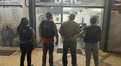 Quatro agentes da Polícia Civil, com coletes da DEIC, escoltam um homem algemado na entrada da Deic.