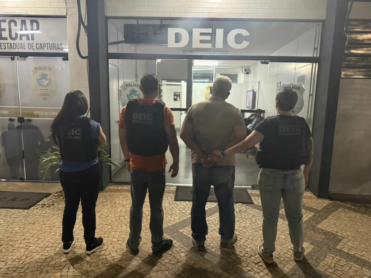 Quatro agentes da Polícia Civil, com coletes da DEIC, escoltam um homem algemado na entrada da Deic.