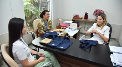 Reunião em gabinete com três mulheres conversando; sobre a mesa, ecobags azul-marinho e pacotes.