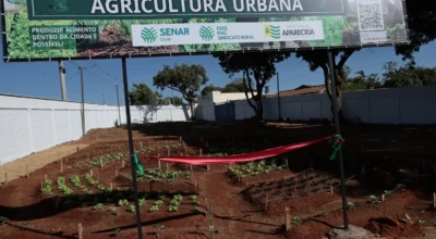 Horta recém-implantada com canteiros e mudas no solo vermelho, cercada por fita de inauguração. Ao fundo, painel “AGRICULTURA URBANA” com logos do Senar Goiás, FAEG e Prefeitura de Aparecida.