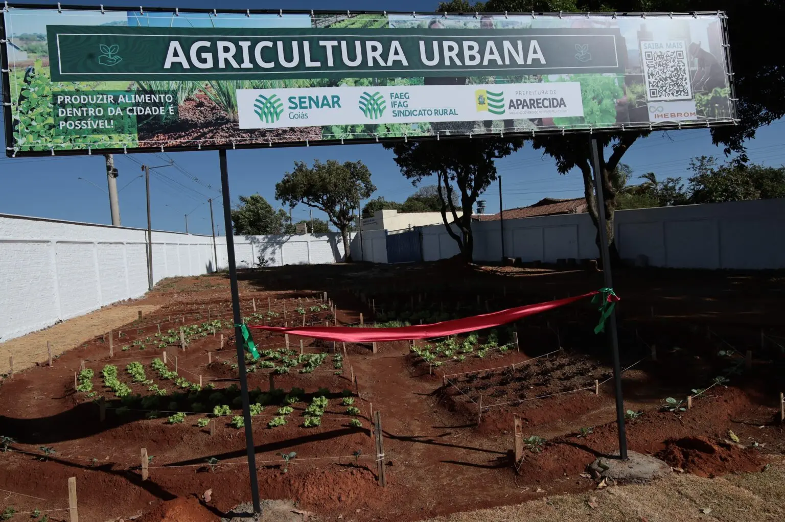 Horta recém-implantada com canteiros e mudas no solo vermelho, cercada por fita de inauguração. Ao fundo, painel “AGRICULTURA URBANA” com logos do Senar Goiás, FAEG e Prefeitura de Aparecida.