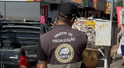 Fiscal da SMTA com colete “Diretor de Fiscalização” em rua comercial de Aparecida. Ao lado, entulho e caçamba ocupam a calçada enquanto pedestres circulam ao fundo.