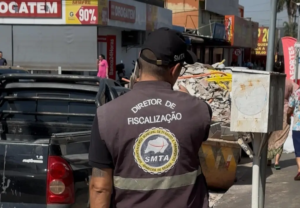 Fiscal da SMTA com colete “Diretor de Fiscalização” em rua comercial de Aparecida. Ao lado, entulho e caçamba ocupam a calçada enquanto pedestres circulam ao fundo.