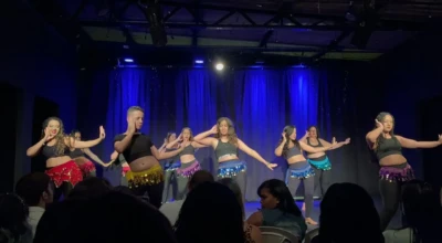 Grupo misto dança dança do ventre no palco, os gestos destacam rosto e quadris, enquanto a plateia acompanha atenta.