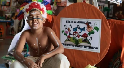Criança sorridente com cocar e pinturas corporais em frente a um estande escolar.