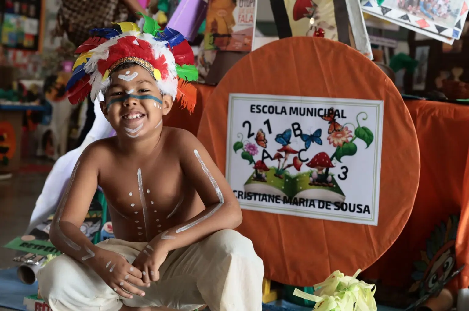 Criança sorridente com cocar e pinturas corporais em frente a um estande escolar.