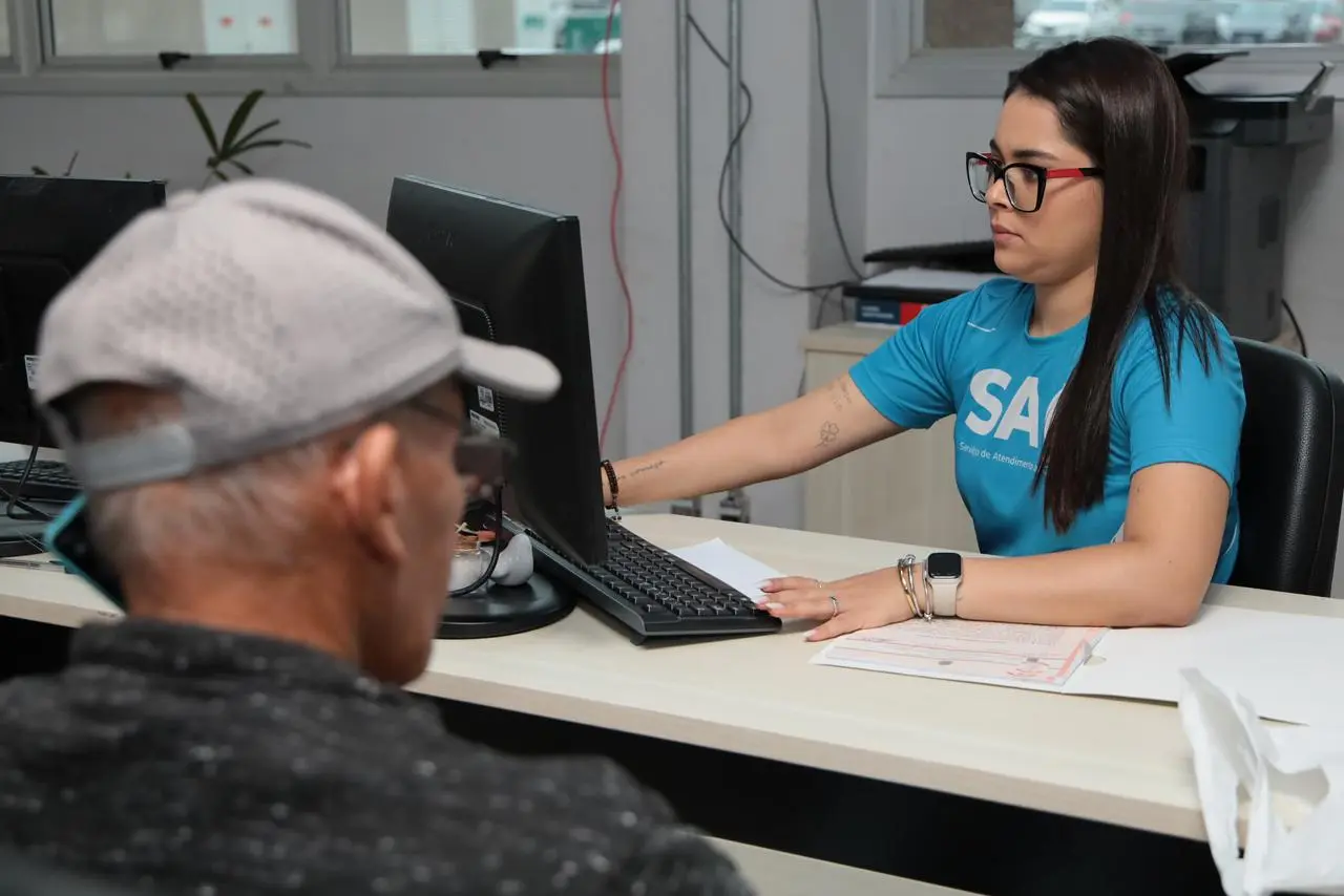Atendente do SAC atende um homem idoso em balcão, usando computador.