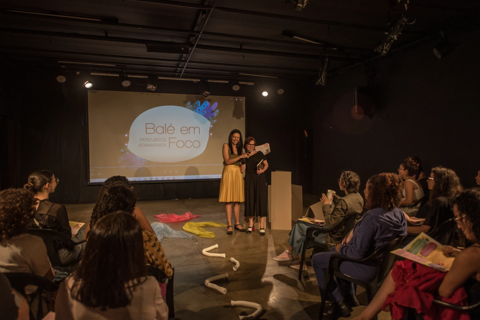 Plateia assiste a duas apresentadoras em palco, diante de telão “Balé em Foco”, com objetos cênicos coloridos no chão.