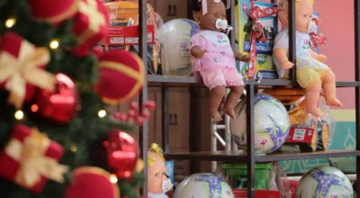 Brinquedos de Natal organizados em prateleiras — bonecas, bolas e jogos — ao lado de uma árvore decorada, prontos para doação.
