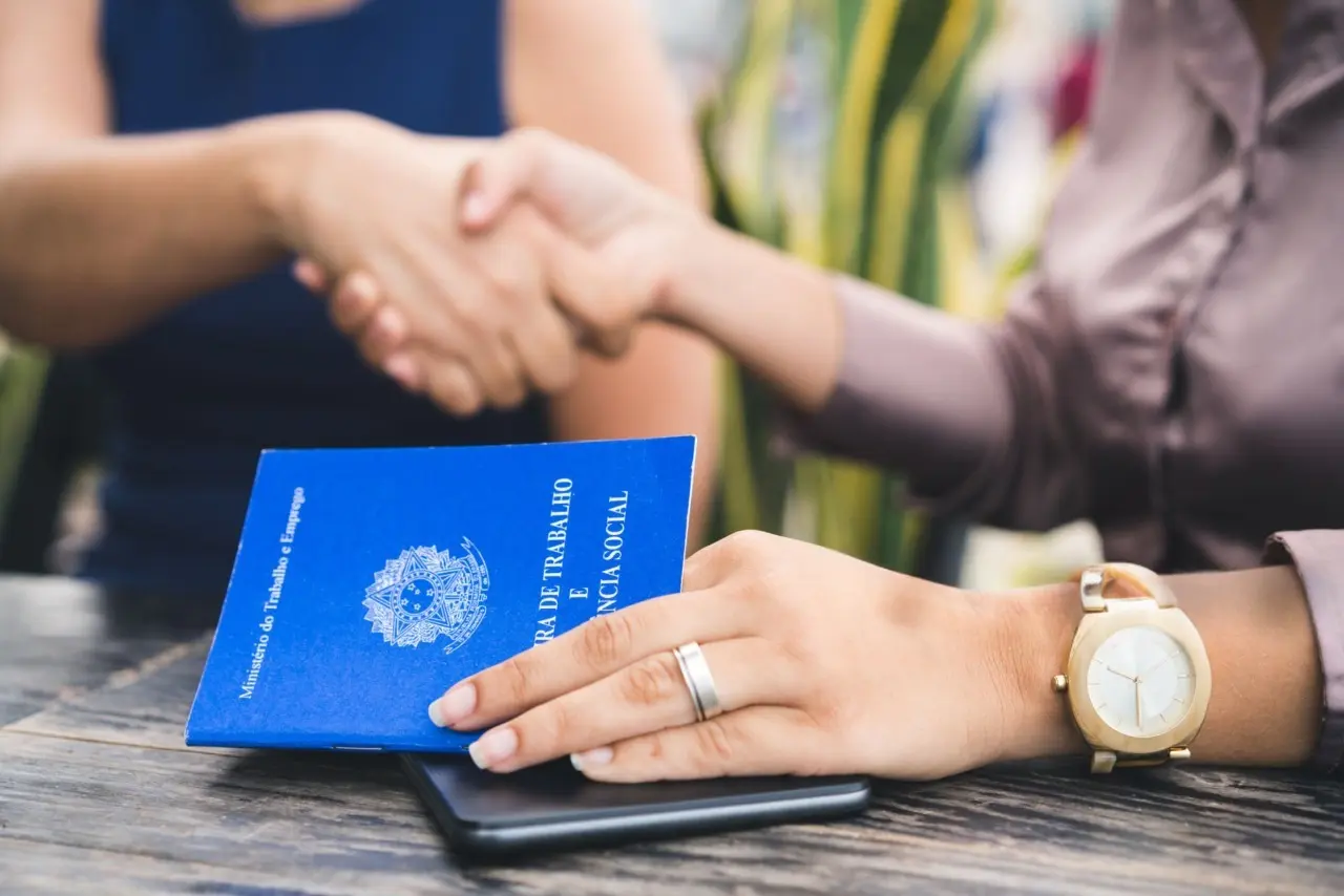 Pessoas apertando as mãos e na mesa uma carteira de trabalho